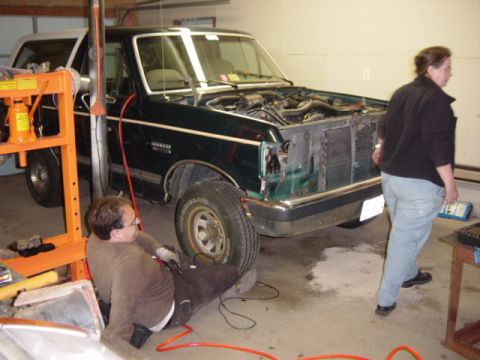 1991 Ford Bronco - A dark green Ford Bronco is parked in a garage with its hood up, as two people attend to it.
