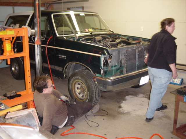 Green SUV with white top in a garage, front end stripped, two people working on the teardown