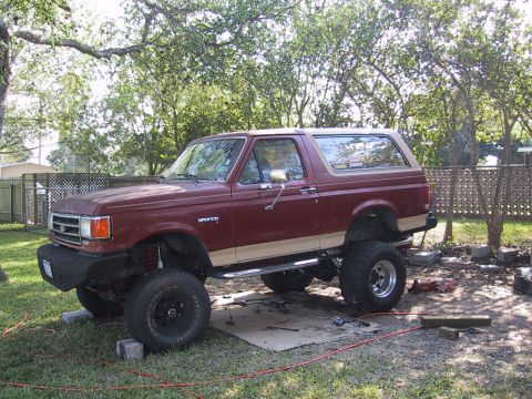 1990 Ford Bronco - This lifted Ford Bronco is parked in a yard with trees and a fence in the background.
