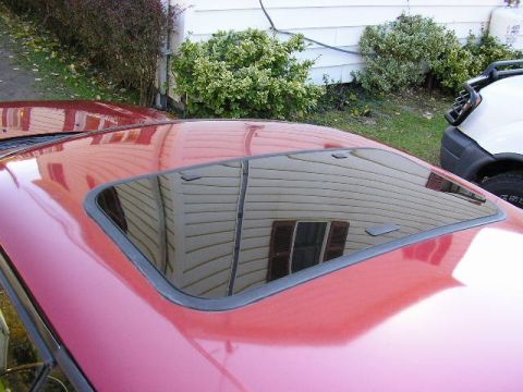 1994 Ford Probe - Close-up of the roof of a red car showing a closed glass sunroof panel, its surface reflecting nearby house siding and shrubs. A white vehi…