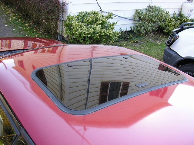 Close-up of a red car roof with a closed glass sunroof reflecting house siding