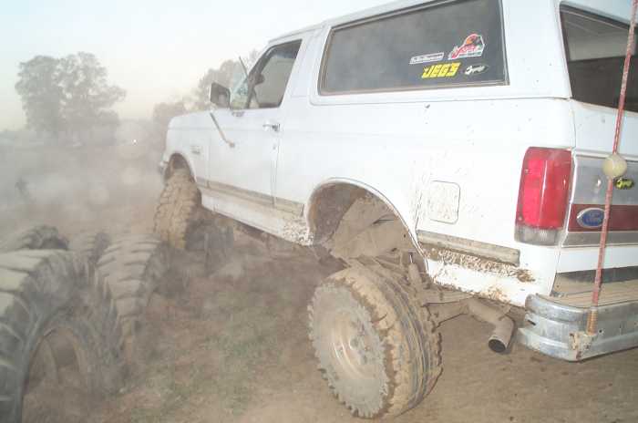 Lifted white Ford Bronco crawling over tractor tires in dust, rear three-quarter view