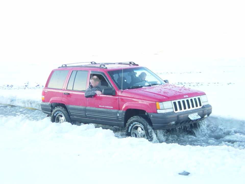 Red Jeep Grand Cherokee stuck in snow, front 3/4 view. Water splashes around tires