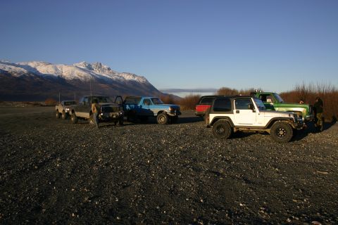 2003 Jeep TJ - A group of off-road vehicles are parked in a lot with mountains in the distance under a clear blue sky.