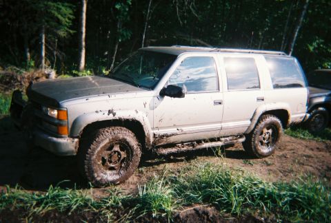 2000 Chevrolet Tahoe - A muddy silver SUV, likely a 1995 model, navigates off-road terrain, with another vehicle partially visible in the background.