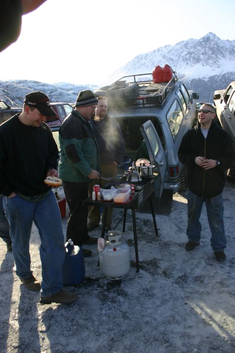 2003 Jeep TJ - People are gathered around a portable grill with propane tank, with vehicles and a snowy mountain range behind them.