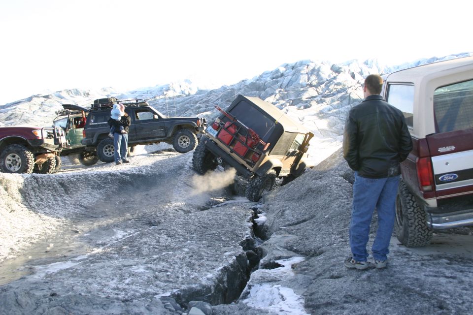 Several off-road vehicles navigate a cracked glacier, with one vehicle tilted precariously