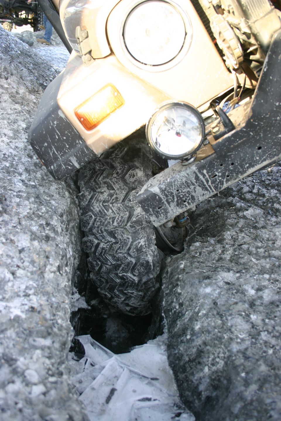 Tan Jeep Wrangler front tire stuck in icy crevice, with auxiliary light visible