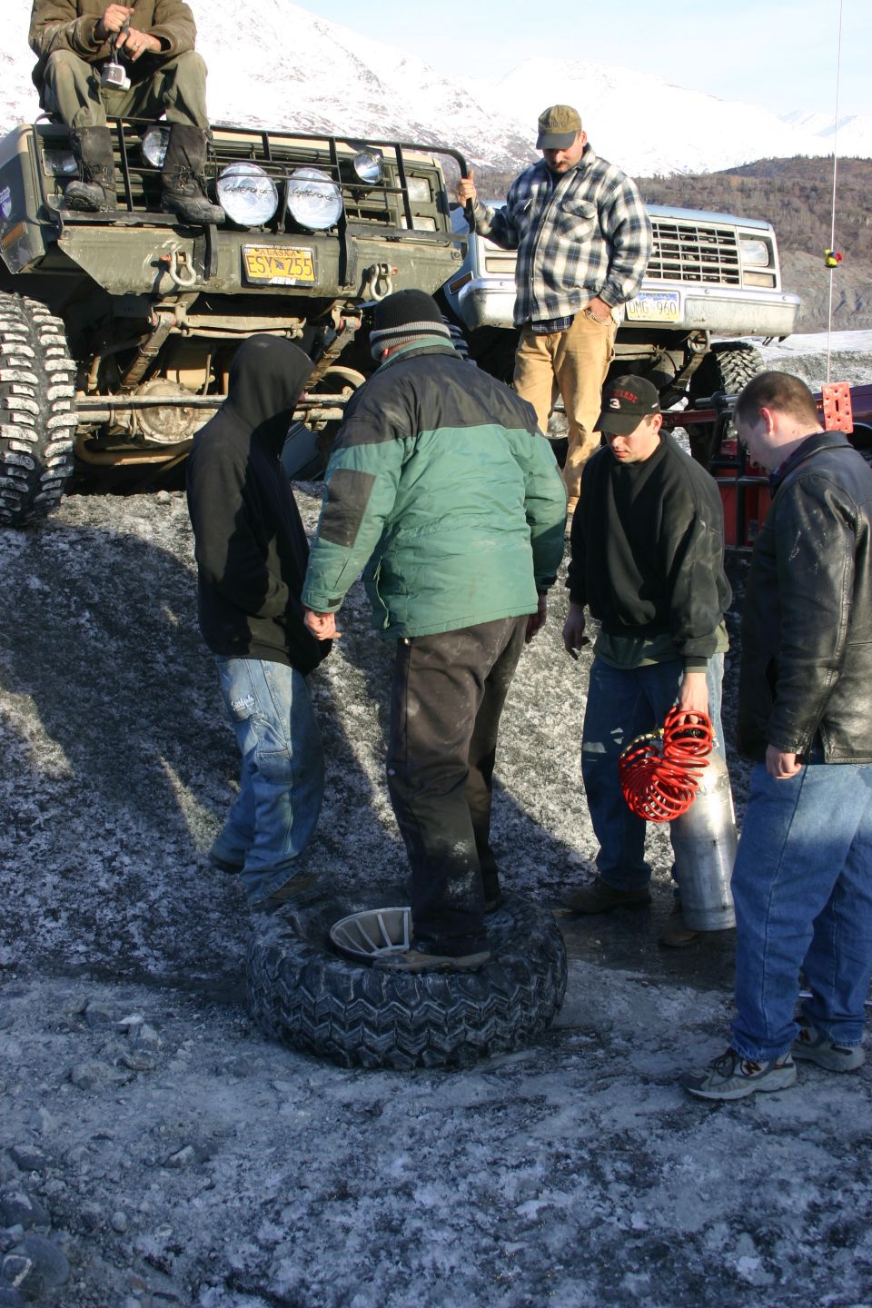 Green Jeep Wrangler with large tires and auxiliary lights, front view. People are gathered around a spare tire on the ground