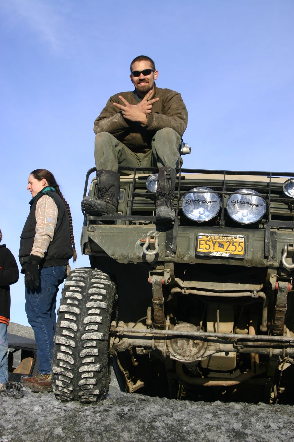 Man wearing sunglasses and a brown jacket sits on the front of a green military-style truck, making a peace sign. Alaska license plate ESY 255 is visible