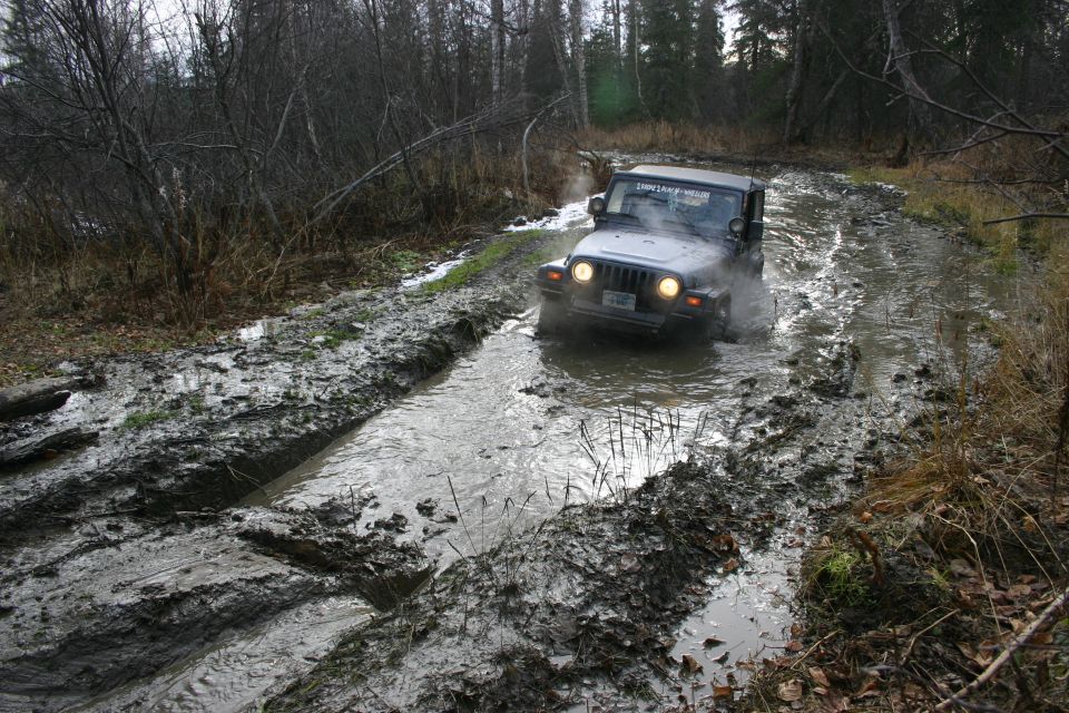 dark gray Jeep TJ drives through a muddy, water-filled trail in a wooded area. The Jeep's headlights are on