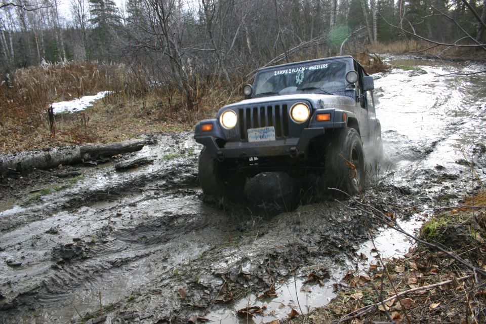 Dark blue Jeep Wrangler TJ driving through mud, front-left quarter view. Headlights on, license plate visible