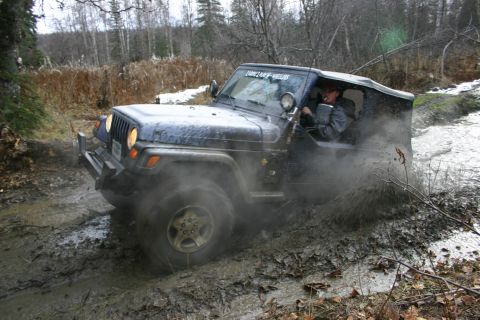 2003 Jeep TJ - A dark blue 2003 Jeep TJ navigates a muddy trail, kicking up spray.