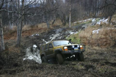 2003 Jeep TJ - A yellow Jeep Cherokee XJ navigates a challenging muddy terrain with trees and sparse vegetation in the background.