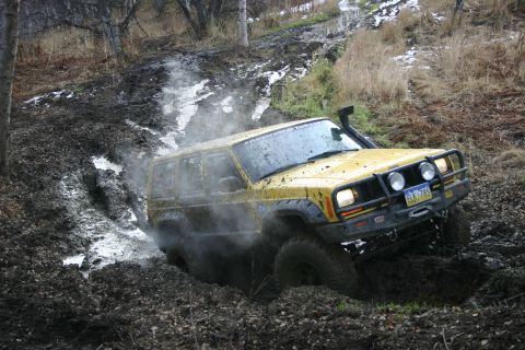 2003 Jeep TJ - A yellow Jeep Cherokee XJ navigates a muddy off-road trail, kicking up spray as it struggles through the terrain.