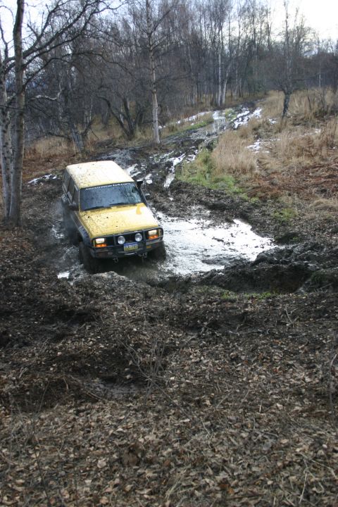 2003 Jeep TJ - A yellow 2003 Jeep TJ navigates a muddy trail in a wooded area, kicking up water and mud.