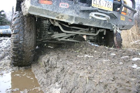 2003 Jeep TJ - A 2003 Jeep TJ's rear axle and suspension are deeply embedded in thick mud, with another vehicle visible in the background.