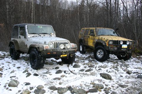 2003 Jeep TJ - Two modified Jeeps are parked on a rocky, snow-dusted surface, surrounded by bare trees.