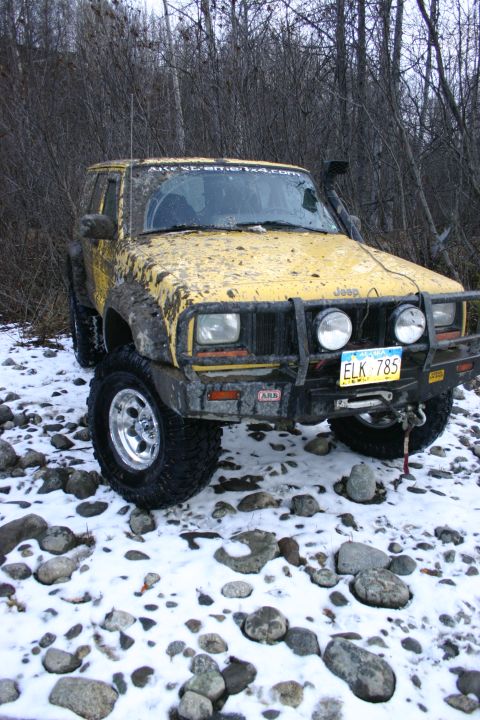 2003 Jeep TJ - A mud-splattered yellow Jeep Cherokee XJ sits on a rocky, snow-covered terrain, equipped for off-road adventures.