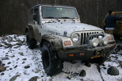 2003 Jeep TJ - A muddy 2003 Jeep TJ Rubicon sits in a snowy, wooded area, with another vehicle visible behind it.