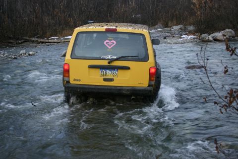 2003 Jeep TJ - Yellow Jeep Cherokee XJ navigates a shallow river, kicking up water.