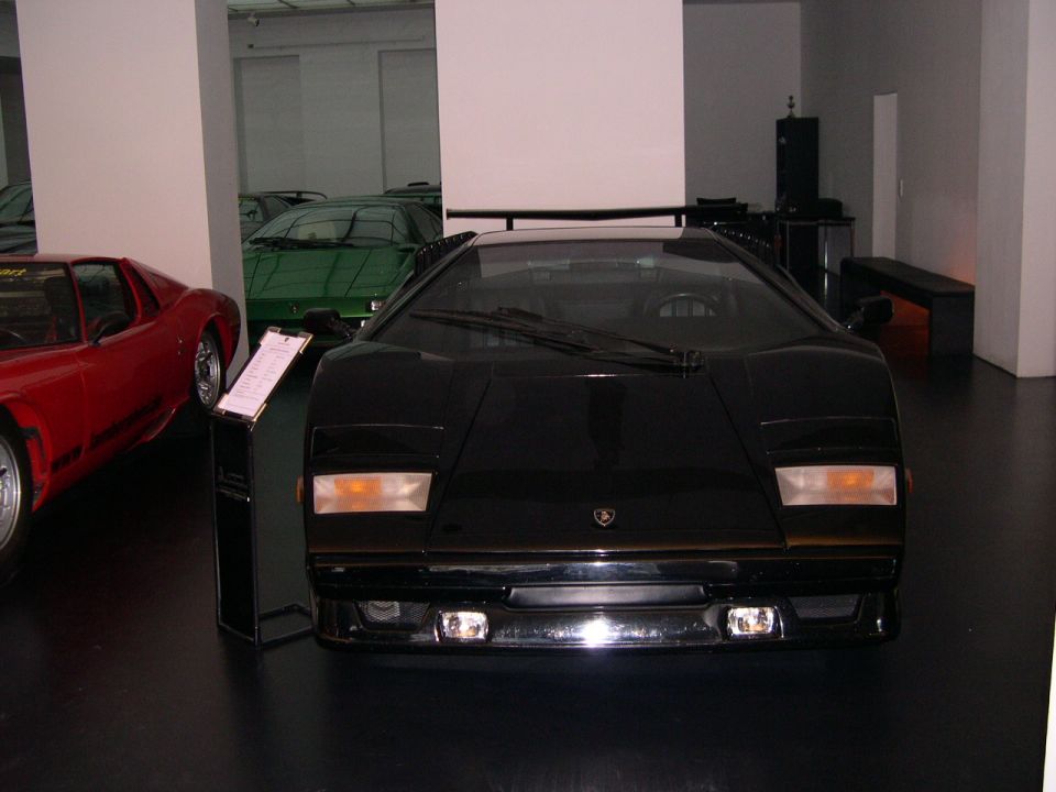 Front view of a black Lamborghini Countach in a showroom with a red car to the left and a green car behind