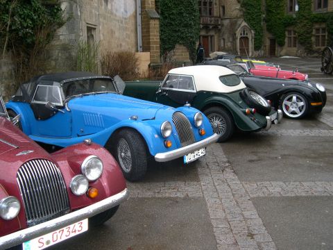 2003 Jeep TJ - A line of classic Morgan cars are parked on a wet cobblestone surface in front of a stone building.