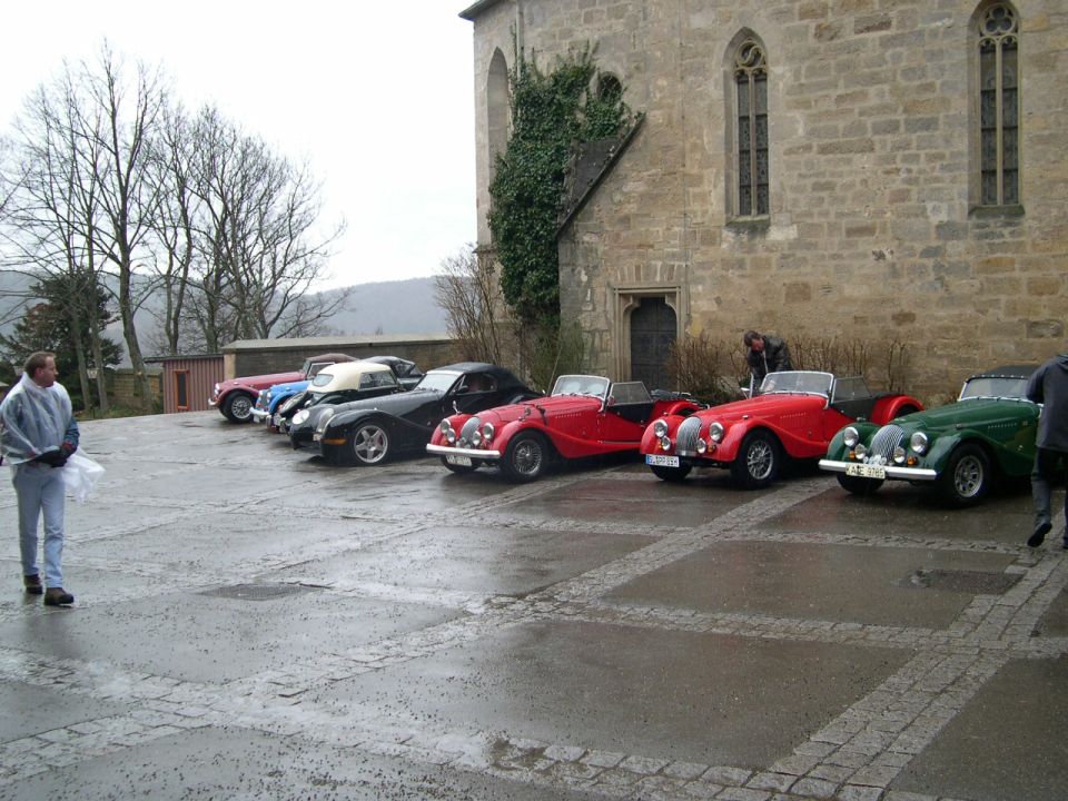 Line of classic red, black, and green sports cars parked on wet cobblestones