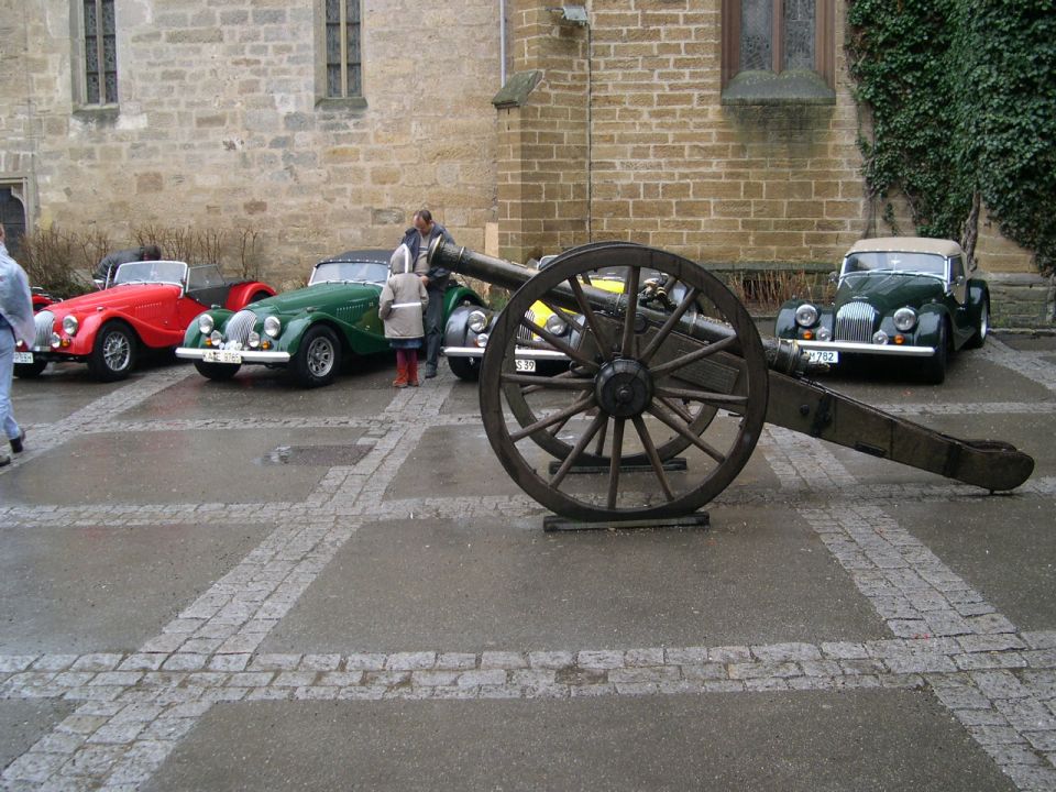 Several vintage convertible cars parked on a cobblestone plaza with a large wooden cannon in the foreground