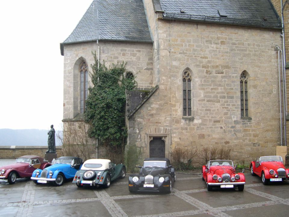 Several classic cars, including a red one and a blue one, are parked in a courtyard in front of a stone building with gothic windows
