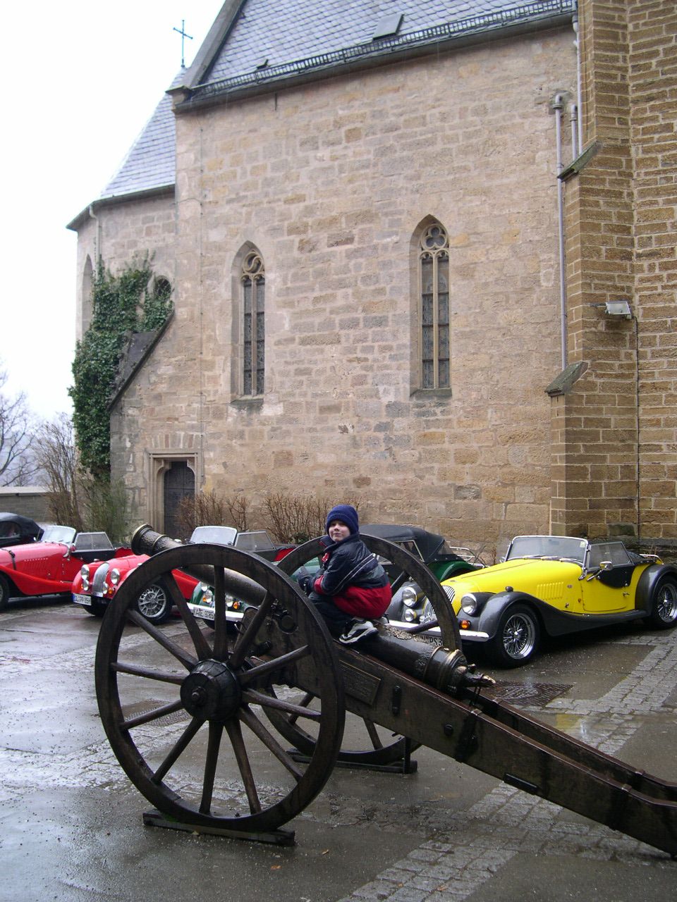 Child sits on a large wooden cannon in front of a stone church. Several vintage cars, including a red one and a yellow convertible, are parked behind the cannon