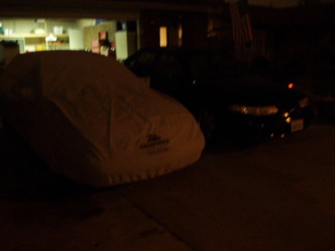 1993 Ford Taurus - A car is covered in a light-colored car cover, with a black sedan parked behind it.