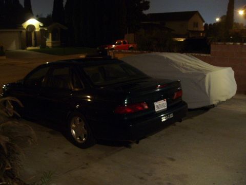 1993 Ford Taurus - A dark green sedan is parked next to a covered vehicle at night, with a red truck in the background.