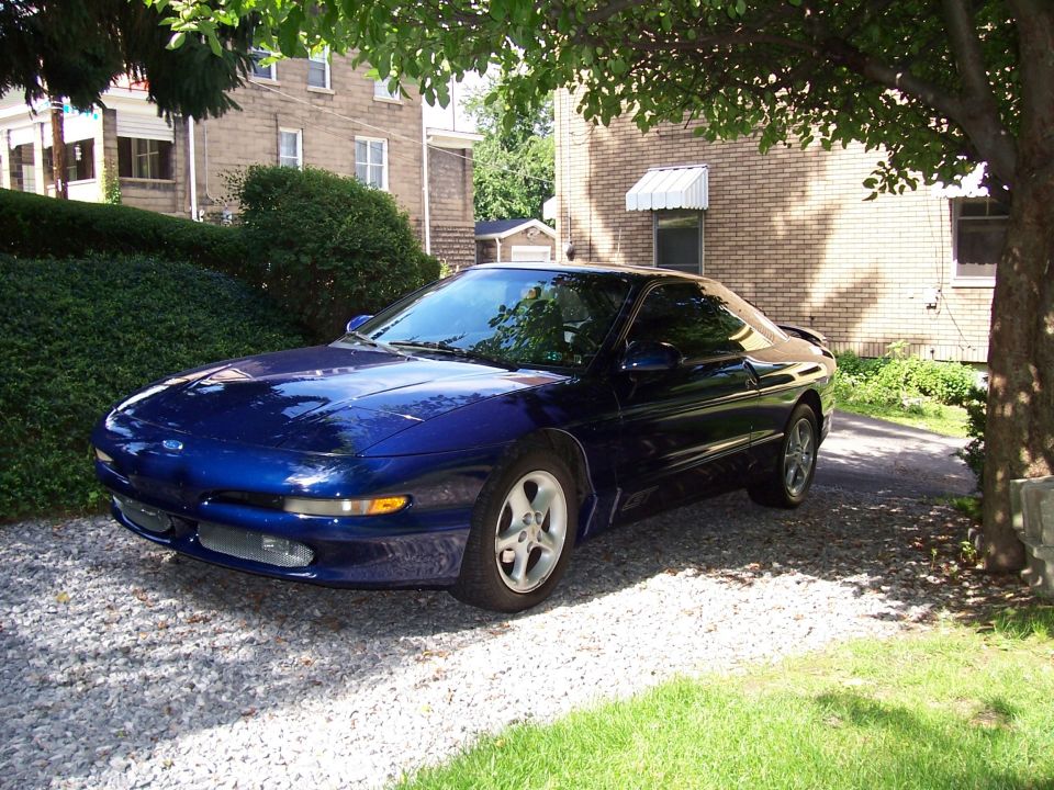 Dark blue Ford Probe coupe parked on gravel driveway, front three-quarter view under a tree