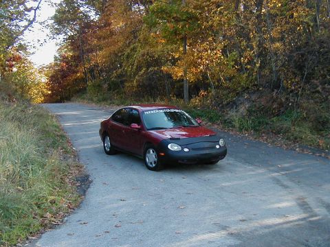 1997 Ford Taurus - A red 1997 Ford Taurus sedan is parked on a winding road surrounded by trees with autumn foliage.