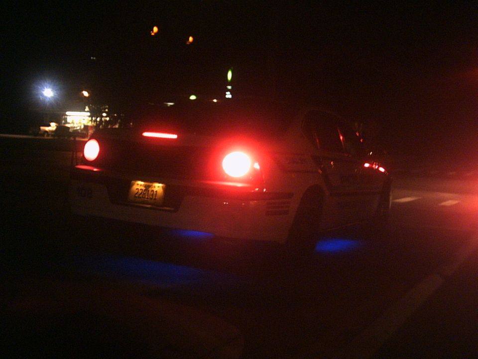 Rear view at night of a light-colored sedan with red taillights lit and blue underglow, yellow plate 228131