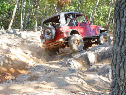 1996 Ford Bronco - A red off-road vehicle tackles a challenging rocky terrain in a wooded area.