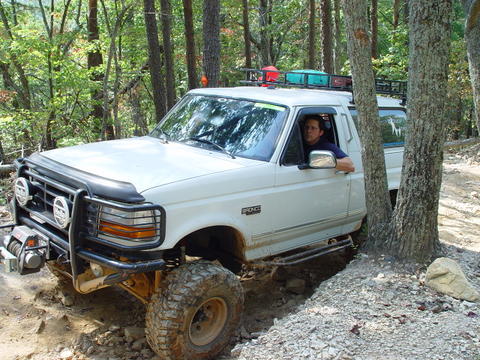 1996 Ford Bronco - A white Ford Bronco navigates a wooded trail with significant off-road enhancements.