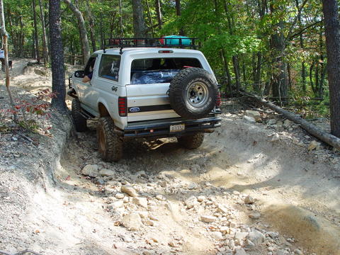 1996 Ford Bronco - A white 1996 Ford Bronco navigates a challenging, rocky off-road trail surrounded by trees.