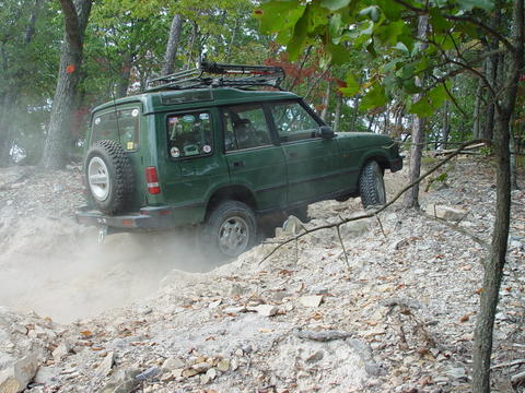 1996 Ford Bronco - A green SUV tackles a challenging off-road trail, kicking up dust on a steep, rocky slope.