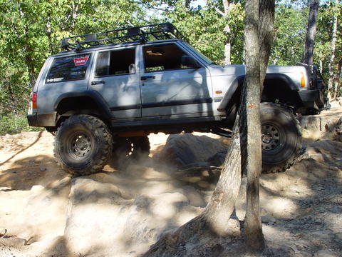 1996 Ford Bronco - A lifted Jeep Cherokee XJ navigates a rocky trail, kicking up dust.