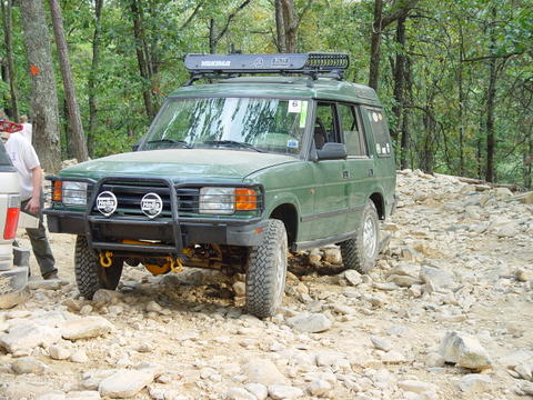 1996 Ford Bronco - A green Land Rover Discovery navigates a challenging rocky path in a wooded area.