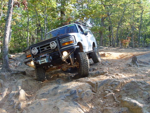 1996 Ford Bronco - A white Ford Bronco navigates a challenging off-road trail with large rocks and trees.