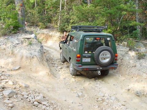 1996 Ford Bronco - A green Land Rover navigates a rocky off-road trail, showing its rear profile and spare tire.