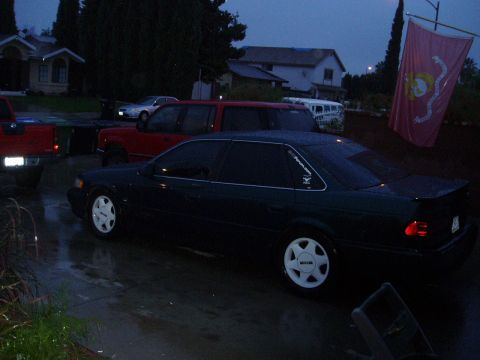 1993 Ford Taurus - A dark sedan with distinctive white wheels is parked on a wet driveway.