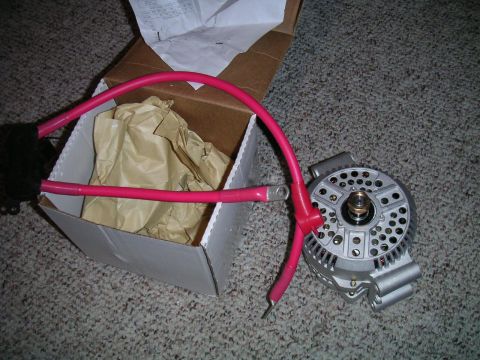 1994 Ford Bronco - New silver alternator laid on carpet next to its opened white shipping box and packing paper, shown face-on with the vented front housing,…