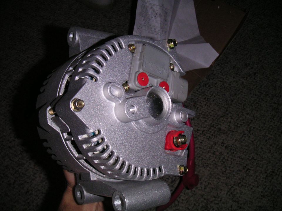 Silver alternator with red B+ cable and voltage regulator held over carpet