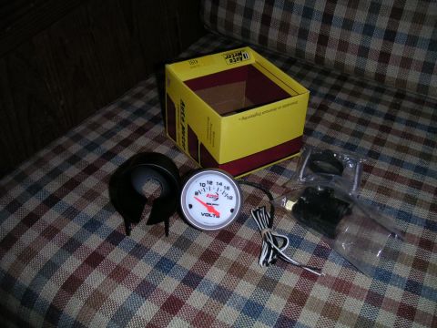 1994 Ford Bronco - An Auto Meter voltmeter gauge and its associated parts are laid out on a patterned surface next to its original packaging.