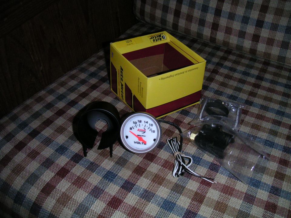 Auto Meter voltmeter gauge, mounting cup, and box on a plaid surface