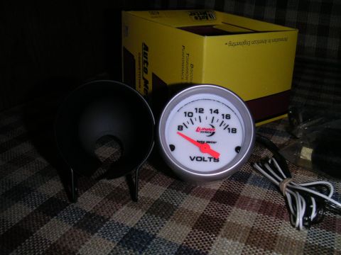 1994 Ford Bronco - Unboxed Auto Meter Lunar Series voltmeter with a white face, red needle and 8-18 VOLTS scale, sitting on a plaid couch next to its black si…